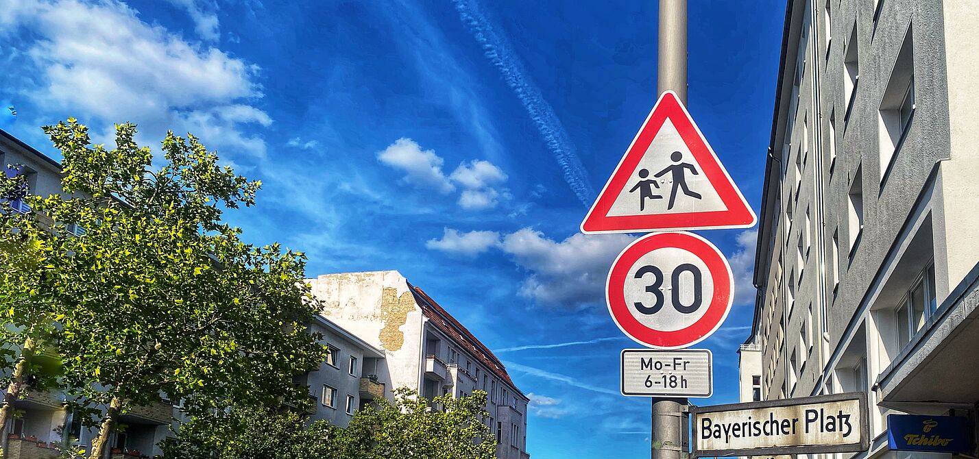 Auf dem Foto sind zwei Verkehrsschilder vor blauem Himmel zu sehen: Tempo 30 und Kinder. Rechts liegt eine Häuserzeile, auf der linken Seite ist die Straße durch Bäume begrünt.