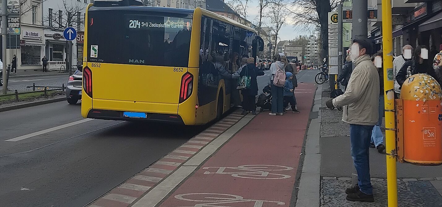 Das Bild zeigt im rechten Bereich eine Haltestelle, daneben ein Haltestellenkap mit darüber geführten Radweg und ein haltender Bus. Fahrgäste steigen ein und aus. 
