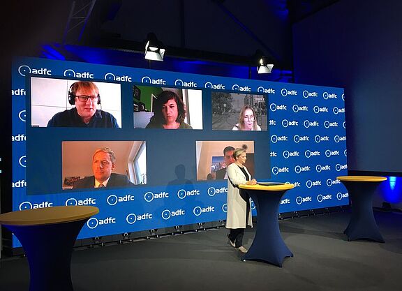 InnoRAD-Symposium Ein Studio mit der Moderatorin am Pult, dahinter eine Videowand mit den Köpfen von per Video zugeschalteten Personen.