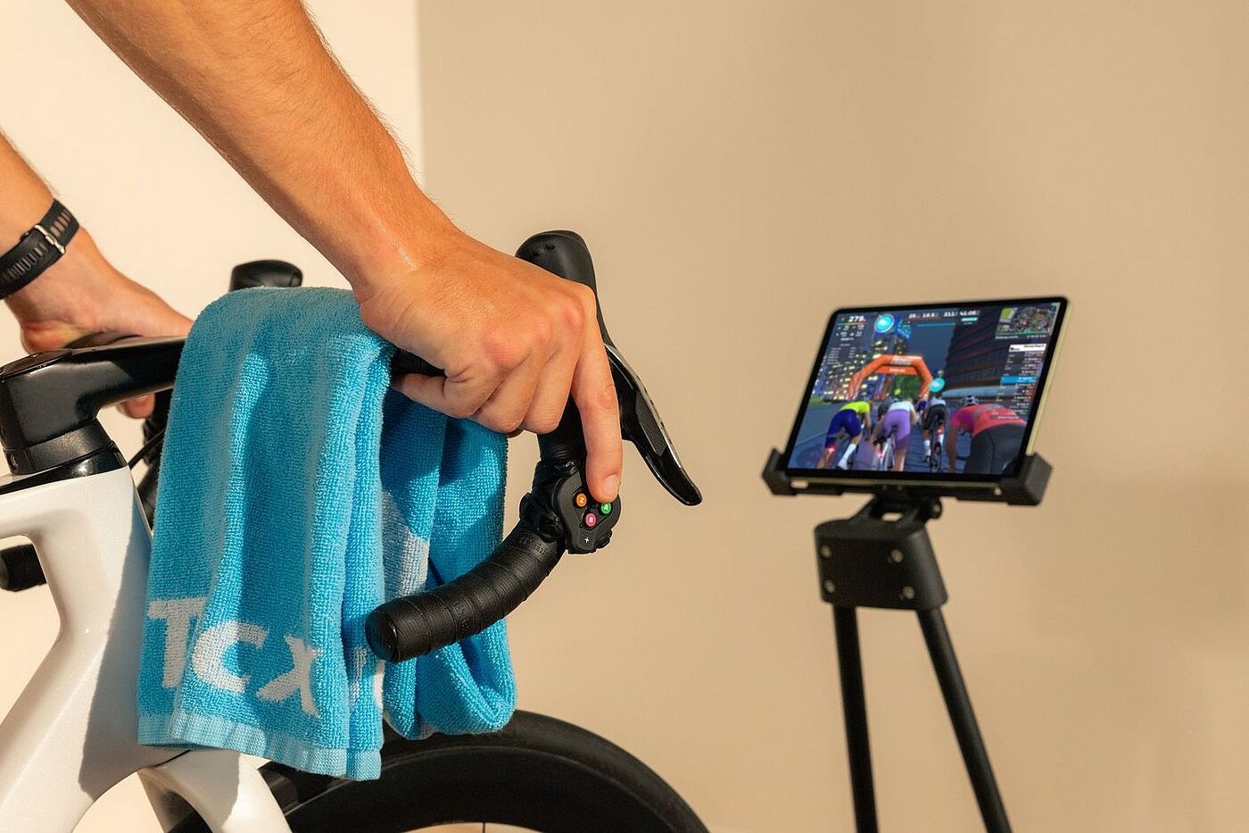 Tacx Neo Tasten Eine Hand hält den Lenker eines Rennrads. Rechts steht ein Tablet auf einem Ständer, auf dem ein Radrennen am Bildschirm zu sehen ist.