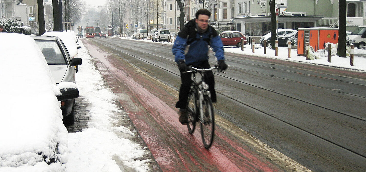 Radfahren im Winter Ein Radfahrer in Winterkleidung fährt auf einem rot markierten Radfahrstreifen neben einer Straße, der weitgehend von Schnee befreit ist, während am Straßenrand und auf geparkten Autos Schnee liegt.